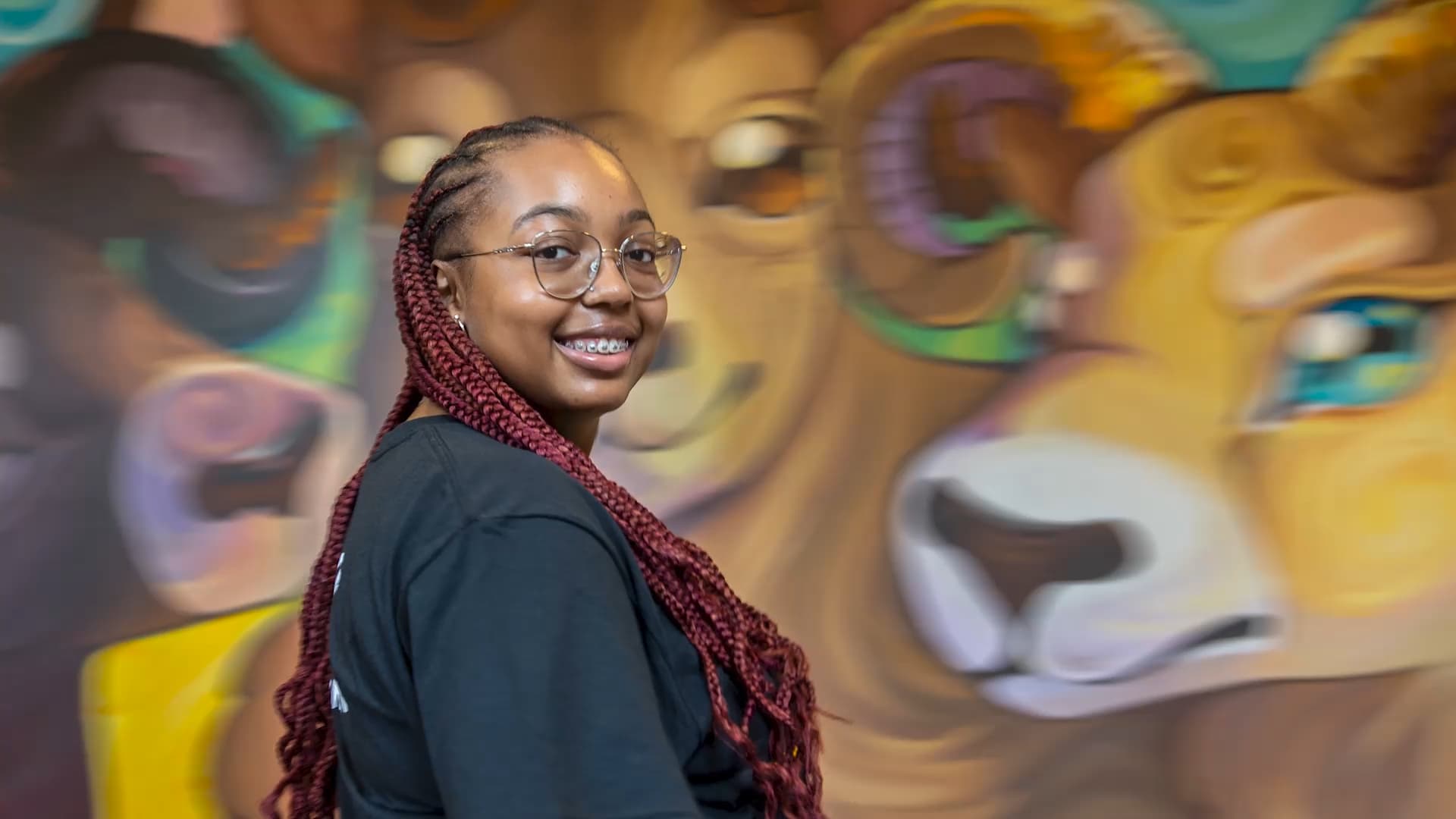 A smiling student standing in front of a whimsical ram mural