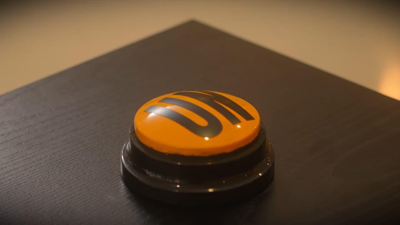 A gold button with the letters UN written in black, sitting on a table waiting to be pressed. What does it do? Go on. Press it.